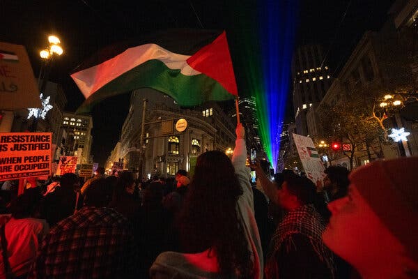 A crowd of people holding signs and a Palestinian flag on a city street at night.